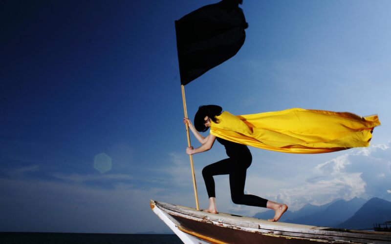 A woman in a bold pose with a black flag and yellow cape on a boat by the sea under blue skies.